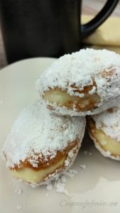 3 Pudding filled donuts coated in powdered sugar on white plate with black coffee mug in background